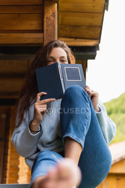 Низький кут веселої жінки сидить на терасі з цікавою книгою і дивиться на камеру під час відпустки в горах Піренеїв. — стокове фото