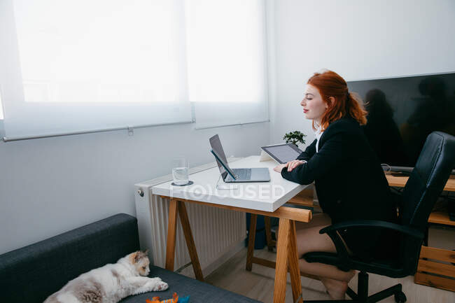Vista lateral de la joven empresaria que trabaja en netbook en el escritorio con tableta y teléfono inteligente en casa - foto de stock