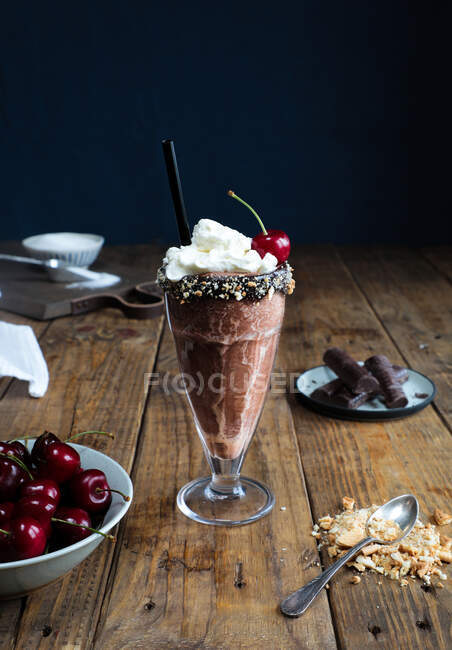 Tazza di frullato al cioccolato condito con panna e ciliegia posta su una superficie di legno su uno sfondo scuro — Foto stock
