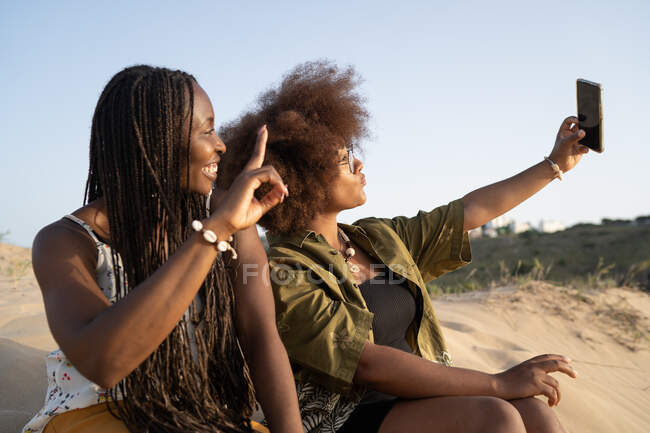 Jovens amigas afro-americanas alegres sorrindo brilhantemente enquanto tomam selfie no smartphone durante o fim de semana de verão na costa arenosa — Fotografia de Stock