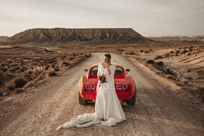 Femme heureuse en robe blanche regardant loin près du véhicule rouge de luxe stationné sur la route poussiéreuse pendant le voyage à travers le parc naturel des Bardenas Reales en Navarre, Espagne — Photo de stock