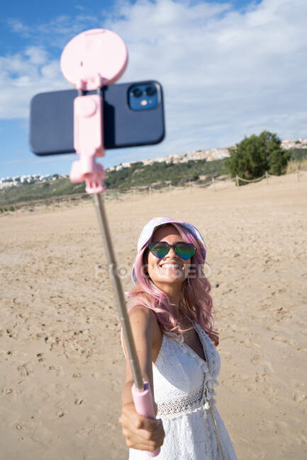 Angle élevé de la femelle ravie aux cheveux roses et en tenue d'été se prendre en photo sur smartphone sur selfie stick tout en se tenant debout sur une journée ensoleillée sur le bord de la mer — Photo de stock