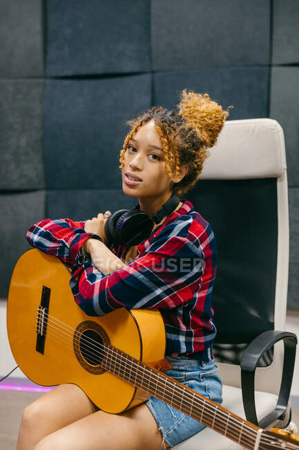 Молодая афроамериканка-гитаристка с классической гитарой и наушниками сидит со скрещенными руками в кресле и смотрит в камеру — стоковое фото