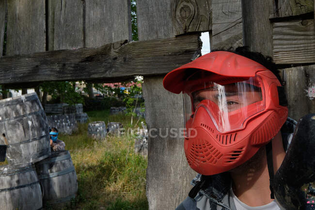 Мужчина в маске и с пейнтбольным пистолетом, стоящим за деревянным забором во время игры — стоковое фото