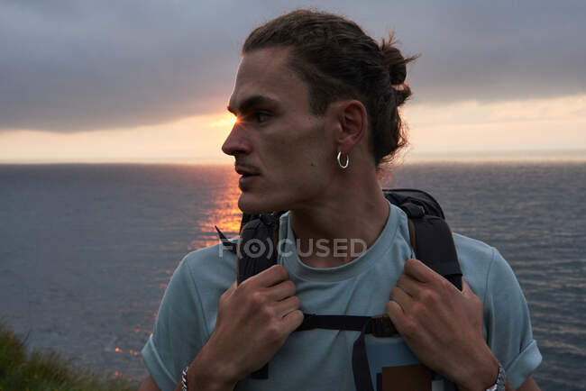 Viajero masculino de pie sobre roca y admirando la vista del mar durante el trekking en verano - foto de stock