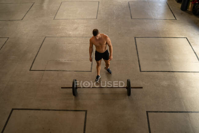 Angle élevé d'un homme athlétique torse nu se préparant pour l'entraînement et s'approchant de haltères lourdes sur le sol dans une salle de gym spacieuse — Photo de stock
