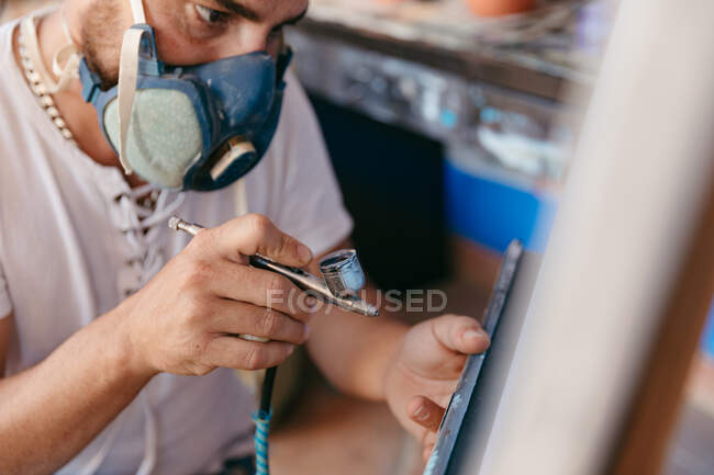 Side view of cropped male artist in respirator using spray gun to paint picture on canvas during work in creative workshop — Stock Photo