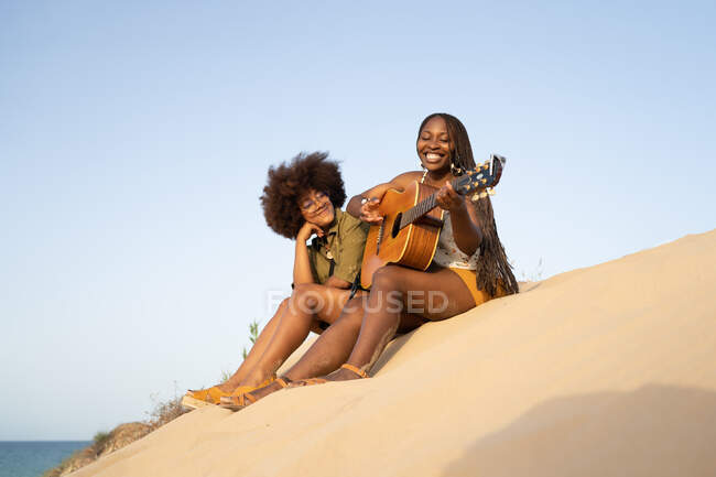 Baixo ângulo comprimento total de jovens amigas afro-americanas felizes tocando guitarra enquanto se sentam juntas na costa arenosa e desfrutando de férias de verão — Fotografia de Stock
