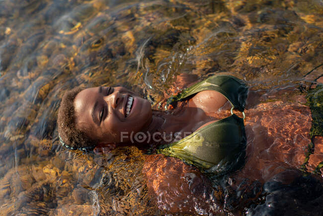 Desde arriba alegre hembra afroamericana sexy en traje de baño acostado con los ojos cerrados en el agua del mar en verano - foto de stock