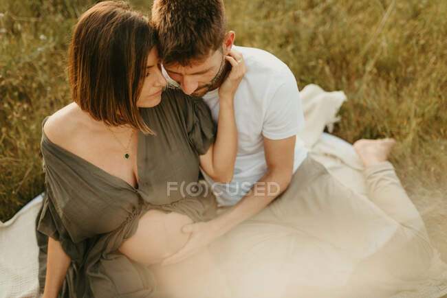 Délicieux câlin masculin de la femelle enceinte avec les yeux fermés tout en étant assis dans la prairie à la campagne — Photo de stock
