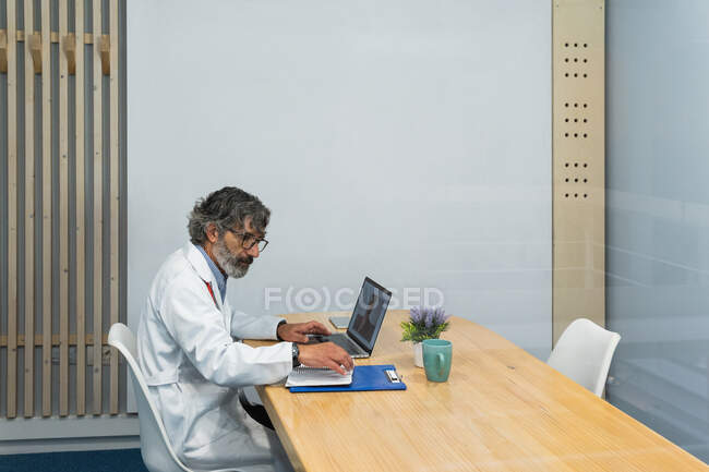 Vista lateral del médico masculino senior que tiene videollamada con el paciente durante la consulta de telemedicina en el hospital - foto de stock