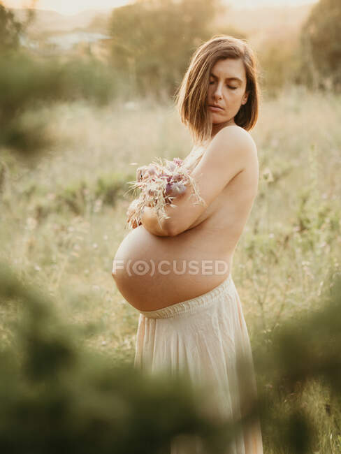 Боковой вид спокойной беременной женщины, обнаженной грудью с букетом цветов, стоящей летом в сельской местности — стоковое фото