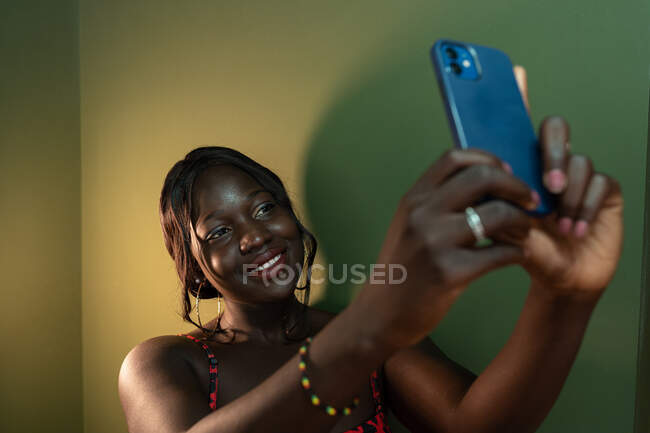 Elegante hembra afroamericana sonriente haciendo un autorretrato con teléfono móvil cerca de la pared en la habitación con poca luz - foto de stock