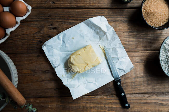 Couteau prêt à couper un morceau de beurre sur une préparation pâtissière sur une table en bois près des œufs et de la cassonade — Photo de stock