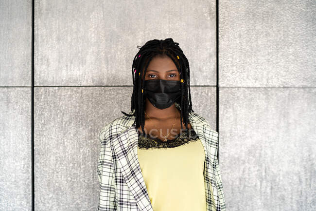 Disappointed African American female in mask standing against gray wall and looking at camera with sadness — Stock Photo