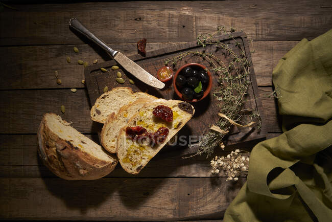 Композиция сверху из нарезанного домашнего круглого хлеба, подаваемого с ароматными сушеными на солнце помидорами на деревянном столе с травами — стоковое фото