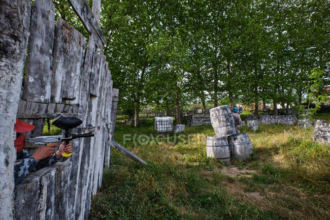 Anonymer männlicher Schütze mit Paintball-Waffe, die auf Feinde abzielt, die sich während des Spiels hinter einem Holzfass verstecken — Stockfoto