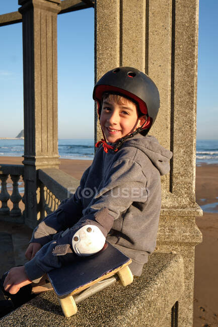 Side view of delighted teenage boy in protective helmet and with skateboard sitting on stone fence near sea and looking at camera — Stock Photo