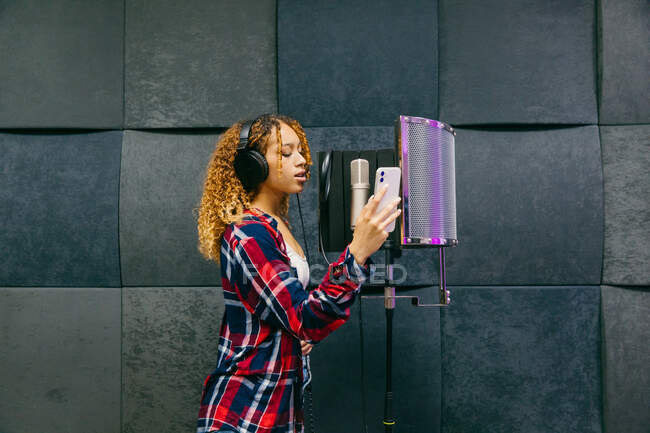 Cantora afro-americana alegre em fones de ouvido com smartphone tocando música contra escudo sonoro em estúdio de gravação — Fotografia de Stock