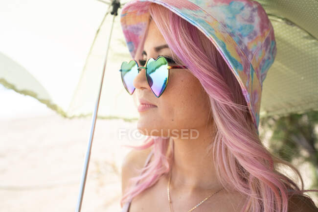 Alto ángulo de mujer alegre con el pelo rosa escondido bajo el paraguas mirando hacia otro lado en la orilla del mar en el día soleado y mirando hacia otro lado mientras disfruta de las vacaciones de verano - foto de stock