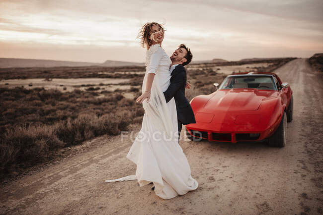 Ganzkörper-Bräutigam umarmt und hebt Braut, während sie am Abend auf der Straße neben rotem Luxusauto im Bardenas Reales Naturpark in Navarra steht, Spanien — Stockfoto
