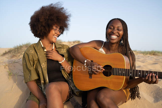 Felices amigas afroamericanas jóvenes tocando la guitarra mientras están sentadas con los ojos cerrados en la orilla del mar y disfrutando de las vacaciones de verano - foto de stock