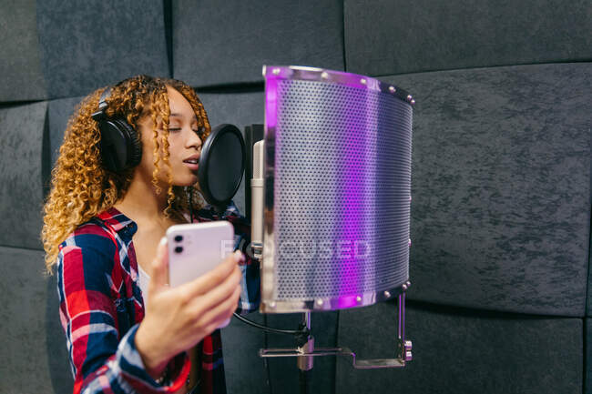 Chanteuse afro-américaine joyeuse dans un casque avec smartphone chantant une chanson contre un bouclier sonore dans un studio d'enregistrement — Photo de stock