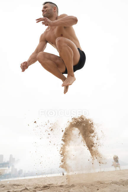 Low angle of male athlete in moment of jumping above sandy seashore during fitness workout in summer — Stock Photo