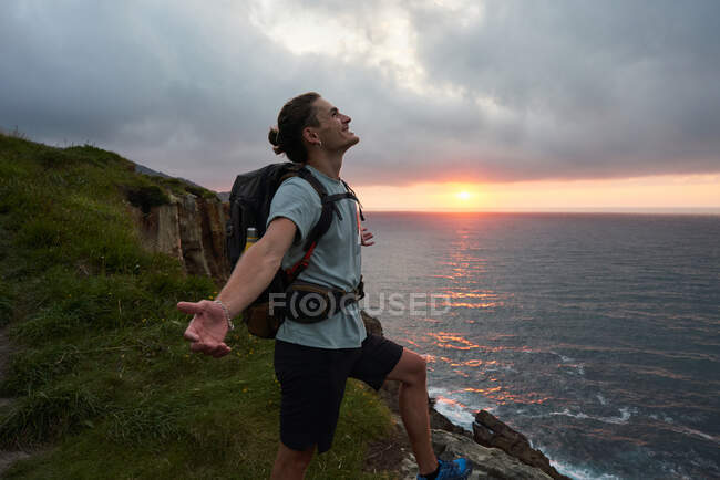 Seitenansicht eines unbeschwerten männlichen Wanderers, der am Aussichtspunkt auf einem Felsen steht und die Freiheit vor dem Hintergrund des Sonnenuntergangs über dem Meer genießt — Stockfoto