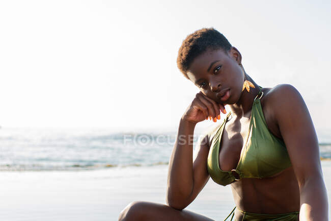 Vista lateral da mulher afro-americana sexy em maiô sentado na água do mar ao pôr do sol no verão — Fotografia de Stock