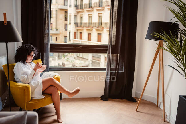 Alto angolo corpo pieno di femmina positiva negli occhiali seduto con le gambe incrociate e mangiare cibo dalla ciotola al mattino — Foto stock