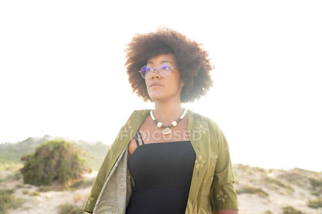 Verträumte junge Afroamerikanerin mit lockigem Haar, die stylische Sommerkleidung mit Halskette und Brille trägt und im Sommer im Sonnenlicht am Strand steht — Stockfoto