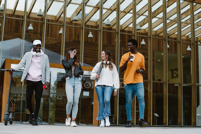 Angolo basso di allegri amici multietnici con smartphone portatili e scooter elettrico che comunicano mentre camminano insieme contro l'ingresso dell'università — Foto stock