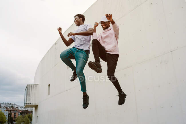 Low angle of full body of energetic African American males in casual wear laughing happily while jumping high together — Stock Photo
