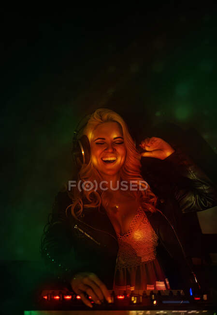 Feliz joven DJ femenino en auriculares sonriendo y utilizando sintetizador para reproducir música mientras está de pie en el humo durante la fiesta en el club nocturno - foto de stock