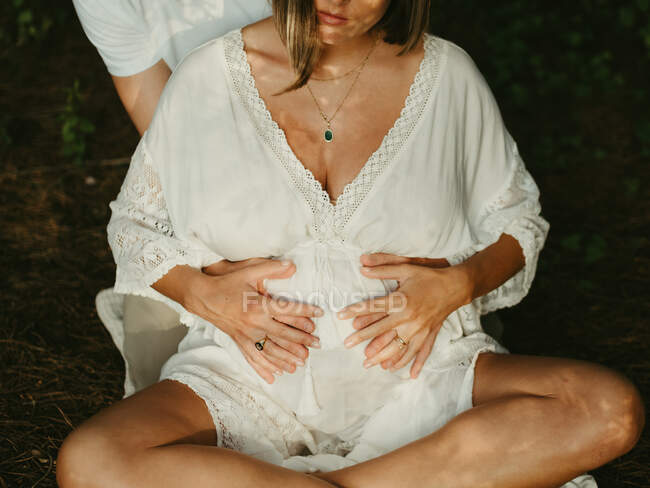 Обрезанный неузнаваемый мужчина обнимает беременную женщину сзади, сидя на сельском лугу — стоковое фото
