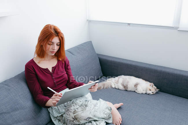 Junge Barfußweibchen mit roten Haaren surfen im Internet auf dem Tablet, während sie mit der Katze auf der Couch zu Hause sitzen — Stockfoto