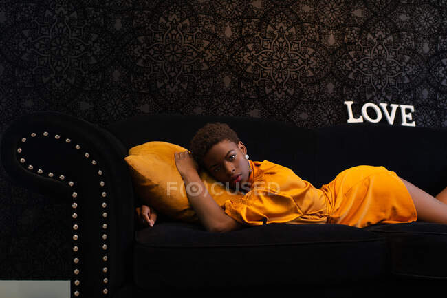Vista lateral de la tranquila mujer afroamericana en vestido naranja brillante tumbado en cómodo sofá negro en habitación oscura y mirando a la cámara - foto de stock