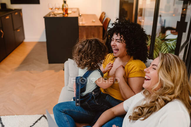 Excited young lesbian couple in casual clothes playing with cute child and sitting together on comfortable couch — Stock Photo