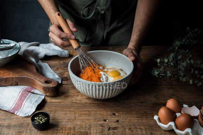 Unrecognizable male cook using whisk to mix pumpkin puree and cream with eggs and flour in bowl while preparing pie on timber table near cutting board and towel — Stock Photo
