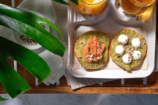De cima de deliciosas torradas de abacate com queijo de salmão e burrata servido na mesa com copos de suco fresco e chá de ervas colocadas perto da planta deliciosa exótica Monstera — Fotografia de Stock