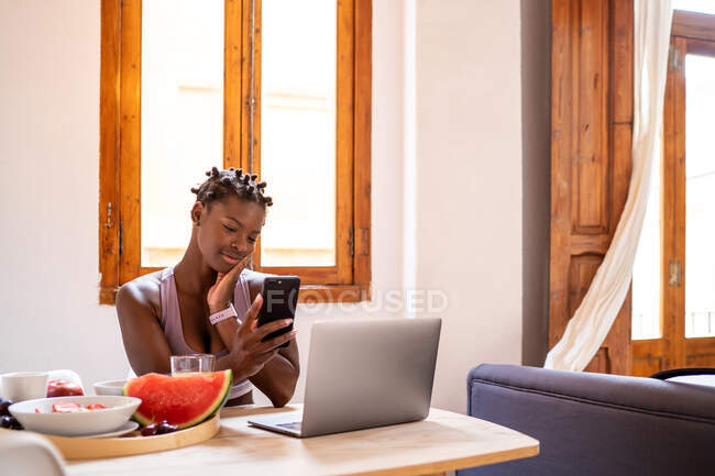 Delighted African American female browsing mobile phone while sitting at table with ripe healthy fruits at home — Stock Photo