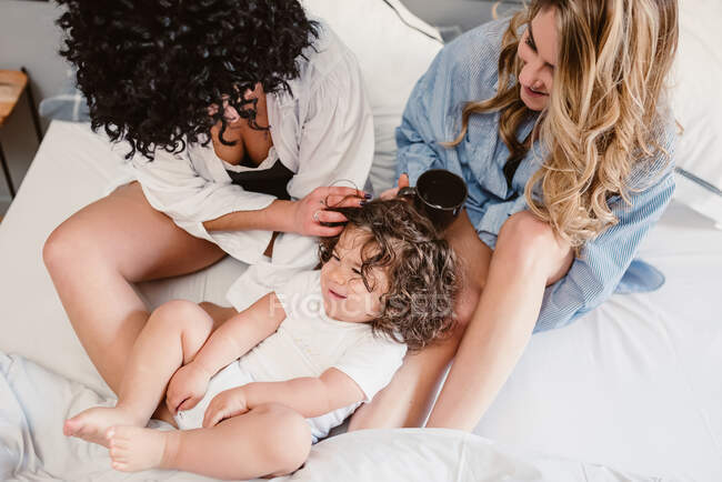 Cultures positives couple de même sexe assis sur le lit et caressant petite fille aux cheveux bouclés dans la chambre — Photo de stock