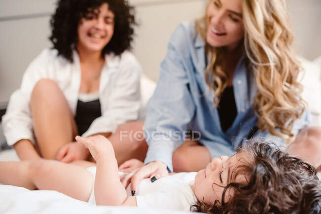 Cultures positives couple de même sexe assis sur le lit et caressant petite fille aux cheveux bouclés dans la chambre — Photo de stock
