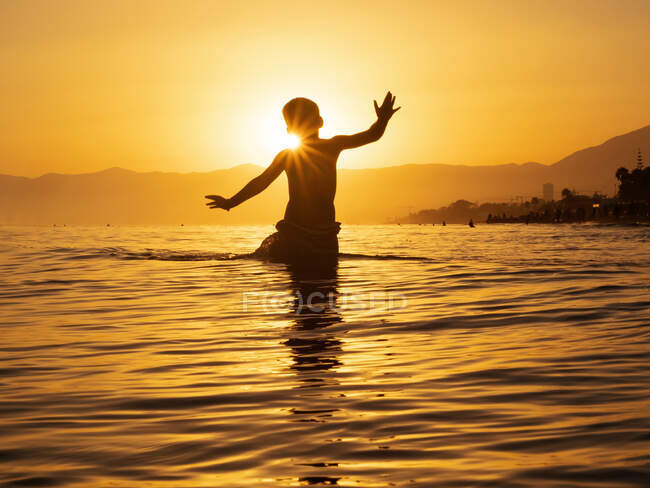 Silhueta de anônimo homenzinho em pé na água do mar contra a luz do pôr do sol no verão — Fotografia de Stock