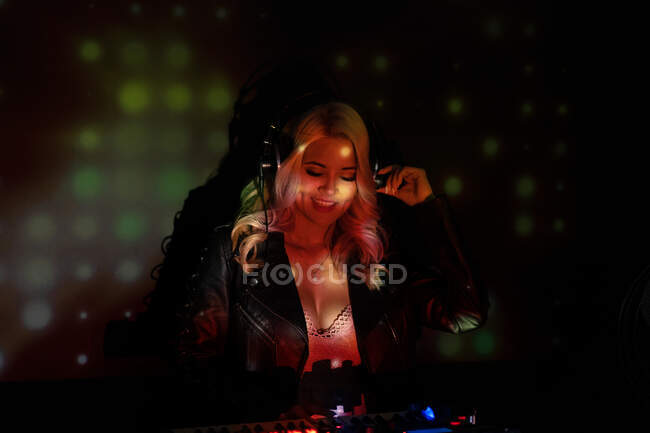 Jovem DJ feliz em fones de ouvido sorrindo e usando sintetizador para tocar música enquanto está de pé em fumaça durante a festa na boate — Fotografia de Stock