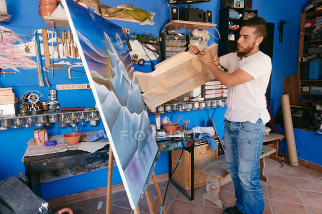 Homem barbudo em roupas casuais removendo papel protetor da tela com paisagem abstrata durante o trabalho no estúdio criativo — Fotografia de Stock