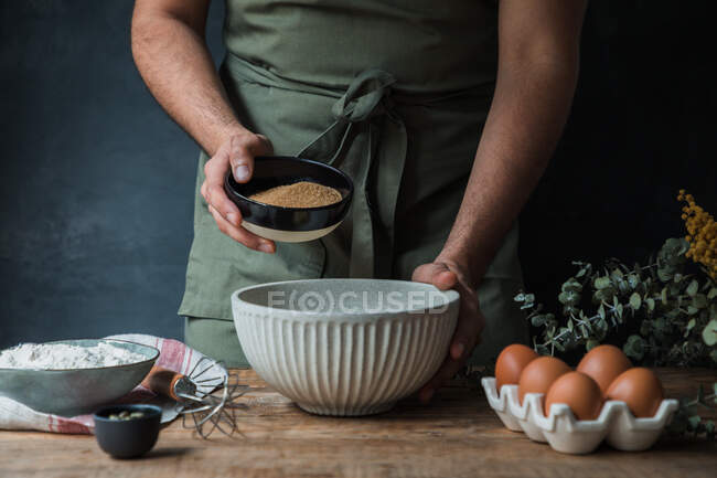 Неузнаваемый человек в фартуке проливает тростниковый сахар в миску возле муки и яиц, готовя тесто для выпечки. — стоковое фото