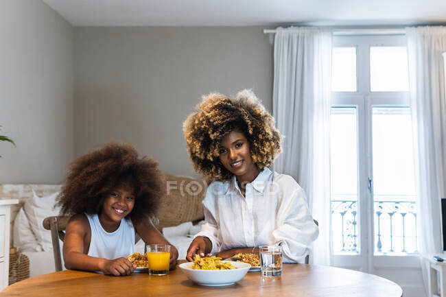 Кудрявые волосы афроамериканской матери и дочери едят аппетитную пищу во время обеда дома и сидя за деревянным столом — стоковое фото