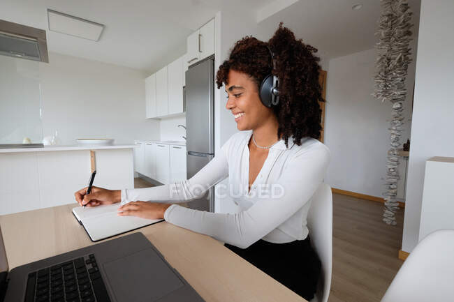 Lächelnde afroamerikanische Freelancerin mit Kopfhörern, die mit Laptop am Tisch sitzt und in Notizblock schreibt, während sie zu Hause ferngesteuert an einem Projekt arbeitet — Stockfoto